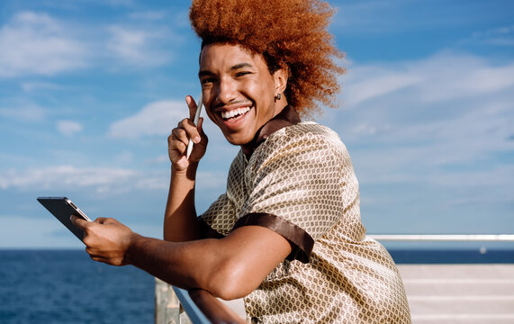 Joyful African American man with curly red afro smiling while using stylus on tablet outdoors, patterned silk shirt, portraying modern technology use, freelance creativity, and innovation.