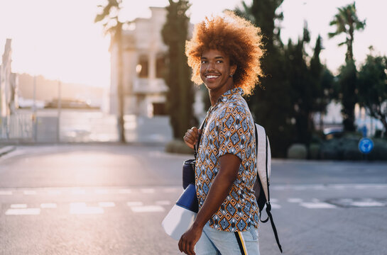 Young man with afro smiling at camera, holding sports gear and backpack. Conceptual urban portrait representing mobility, fitness, individuality, and technology-driven active lifestyles.