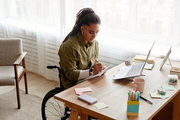 Young adult woman with disability sitting in wheelchair writing in notebook at desk using laptop computer, working in modern office environment with organized workspace