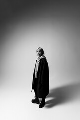 A woman stands in a stark, minimalist setting, dressed in a long coat and comfortable footwear. Her thoughtful gaze and relaxed stance convey a blend of confidence and ease