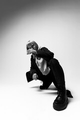 A stylish individual squats in a sleek studio, wearing bold black boots and an oversized outfit while holding a glass. Their expression conveys a blend of sophistication and ease