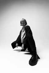 A young individual poses in a crouched position, wearing a grey hoodie and black pants. The minimalist studio environment highlights the fashion and expressive stance, reflecting confidence and style