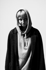 A confident young woman stands against a plain backdrop, wearing an oversized coat over her hoodie. Her expression is bold, showcasing her unique style in an artistic manner