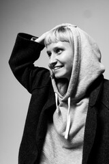 A young woman stands in a cozy, simple space, her short hair complementing her playful expression. She wears a gray hoodie under a dark coat, creating a relaxed vibe