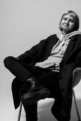 A relaxed person sits comfortably in a chair, wearing a cozy hoodie and a long coat. Their thoughtful expression and casual attire create a laid-back atmosphere in the studio