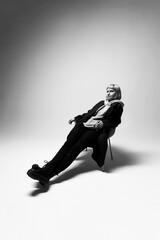 A fashionable young woman leans back casually on a chair, dressed in all black. The soft lighting creates a striking contrast, enhancing her relaxed yet confident pose