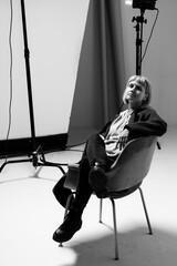 A young person leans back in a modern chair, posing casually during a photoshoot in a minimalist studio setting. Soft lighting adds a dramatic effect, creating an artistic atmosphere