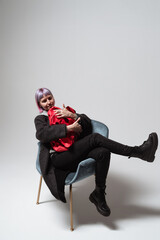 A cheerful woman sitting comfortably in a chic chair embraces a bright red blanket. She has short purple hair and wears a fashionable outfit, creating a lively atmosphere in a minimalist space