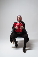 In a bright studio, an individual sits confidently on a chair, cradling a large red gift. Their expressive look and unique style enhance the festive atmosphere of the scene