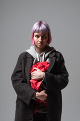 A young woman stands in a minimalist studio, her dark coat contrasting with her pastel pink hair. She clutches red paper tightly, revealing complex feelings in her expression