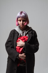 In a minimalist studio setting, a person with pastel hair gently clutches a bundle of vibrant red fabric, their expression reflecting deep emotion