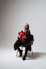 A person with pastel hair smiles brightly while holding a striking red gift close. Seated on a modern chair in a simple setting, the scene captures pure happiness and excitement