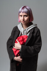 In a modern studio, a young woman with pastel purple hair wears a dark coat over a gray hoodie. She tightly clutches vibrant red gift wrap, looking thoughtfully into the distance