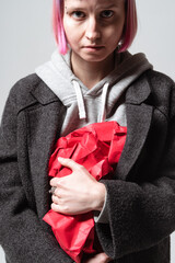 A person stands against a simple background, holding a crumpled red paper tightly. Their expression is serious, and their pink hair pops against the gray coat they wear