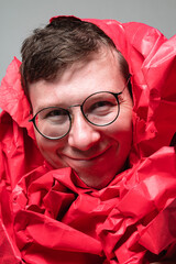 In a colorful studio, a young man joyfully poses with his face wrapped in vibrant red paper. His playful expression captures the essence of fun and creativity in this lively moment