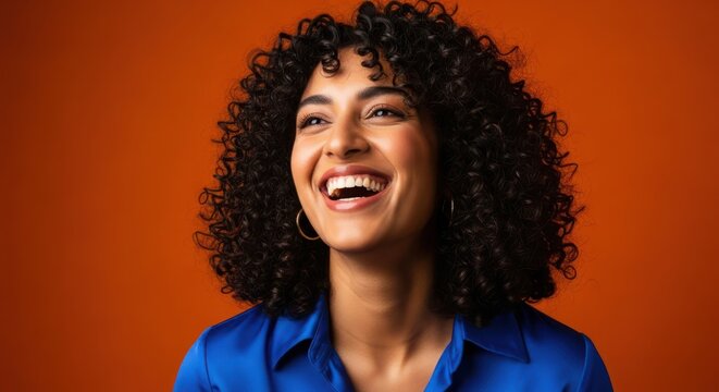 Portrait of a cheerful young indian woman with curly hair, looking up and laughing, isolated on an orange background, full of joy - Powered by Adobe