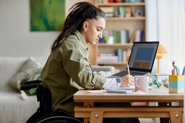 Young adult woman with disability sitting in wheelchair working at desk using laptop and writing in...