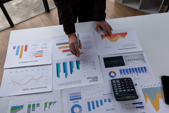 Business professionals collaborate at a desk, analyzing financial reports and data on a laptop. They use charts and graphs to discuss trends and insights in a modern office setting
