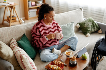 Young adult woman sitting cross legged on sofa holding mug and looking at laptop screen with wheelchair visible beside couch, breakfast food and snacks arranged on coffee table