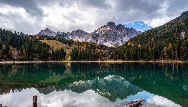 A tranquil alpine lake mirrors a dramatic mountain range and a forest displaying autumn colors, under a dramatic, cloudy sky.