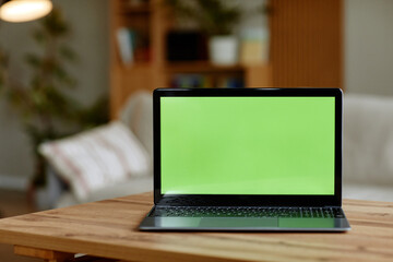 Laptop computer standing on wooden table with green screen display in modern living room, background showing blurred sofa and indoor plants, technology device ready for content insertion
