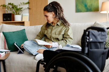 Young adult woman with disability sitting on sofa reading book and using digital tablet, wheelchair visible in foreground, looking away from device, casual home setting