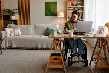 Young adult Caucasian woman with disability sitting in wheelchair working at desk with laptop and...