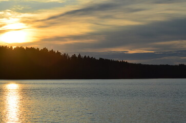 A large lake at sunset. A sunny evening. The sky is covered in patches of cloud, through which the sun shines. A wide lake with slight ripples on the surface reflecting the sun's rays.