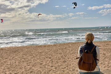 A woman with a backpack sits on a beach and looks at the sea where kitesurfers glide over the waves under a bright sky. beach leisure