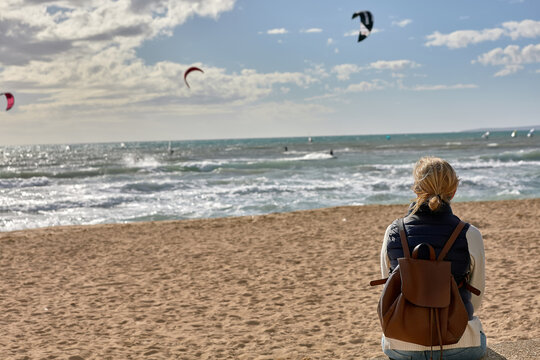 A woman with a backpack sits on a beach and looks at the sea where kitesurfers glide over the waves under a bright sky. beach leisure - Powered by Adobe
