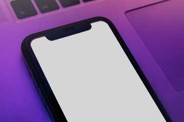 Close-up of smartphone with blank white screen lying on laptop under vibrant pink and purple neon illumination, mockup template for application presentation. Copy space