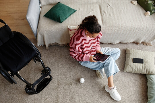 Young adult woman with disability sitting on carpet using digital tablet near wheelchair in home setting, holding device with one hand, looking at screen, relaxed posture - Powered by Adobe