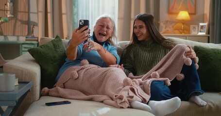 Grandmother and granddaughter laughing together while using funny mobile apps on a smartphone at home, enjoying family bonding and playful technology moments on the living-room sofa.
