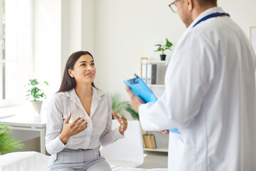 Young cheerful woman patient sitting on couch in medical office talking with male doctor therapist holding report file with appointment and giving consultation during examination in clinic.