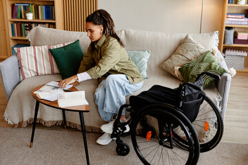 Young adult woman with disability sitting on sofa sorting through paperwork on small table, wheelchair positioned nearby, focused on organizing documents in living room setting