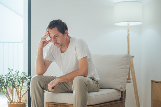Portrait of sad depressed man sitting alone in living room, looking down with regret and emotional pain in quiet low-light home environment.