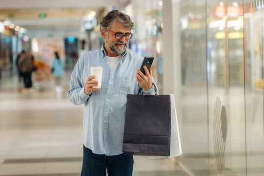 Cup of drink and smartphone are in hands. Senior man is in the mall, shopping time conception - Powered by Adobe