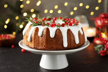 Tasty Christmas cake with icing, cranberries and rosemary on black table against blurred lights, closeup