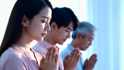 A multi-generational Asian family praying together indoors. A young woman, man, and senior father with hands together in a gesture of faith and respect. Spirituality and tradition concept - Powered by Adobe