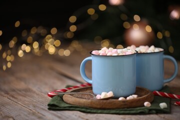 Tasty cocoa with marshmallows and candy canes on wooden table against blurred Christmas lights, closeup. Space for text