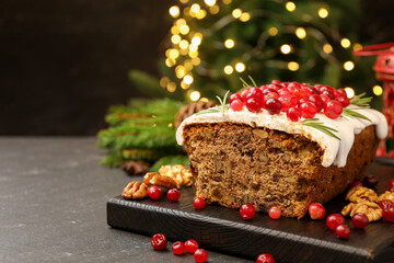 Tasty Christmas cake with icing, cranberries, nuts and festive decor on black table against blurred lights, space for text. Bokeh effect
