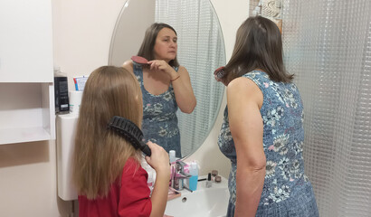 Woman and girl brushing hair together in bathroom mirror  