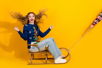 Fototapeta premium Joyful young woman celebrating Christmas on a sled with festive decorations against a vibrant yellow background
