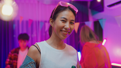Diverse multi ethnic young adult Asian lady wear futuristic neon glowing glasses smile and drink alcohol beer fun enjoy hangout party in living room night indoor home. Disco lights illuminating house.