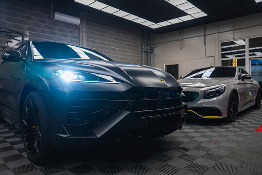 A matte black Lamborghini Urus with one LED headlight on stands in a modern workshop, beside a silver Mercedes AMG coupe with neon yellow accents, under cool panel lights.
