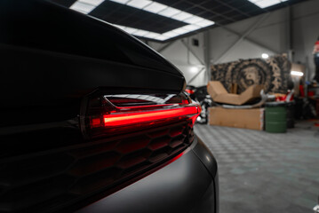 A close up shows the left rear LED taillight glowing on a black luxury SUV inside a high end workshop with skylights, tools, large wheels, and boxes in low key light.