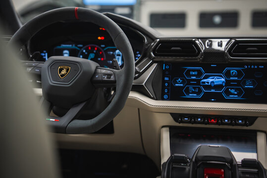 Close view of a Lamborghini Urus cockpit in a high end workshop, cream and black leather, red mode accents, digital gauges, twin screens, and fighter jet style start switch.