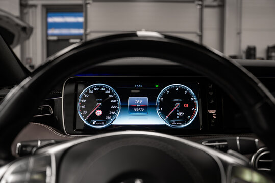 Driver's eye view shows twin gauges and steering wheel, speed and rpm flanking data screen, in a black dashboard within an upscale workshop under bright indoor light