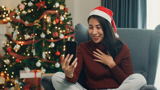 Happy Asia girl wear red santa hat new normal celebrate christmas party at home with smartphone online virtual event video call conference with friend an family on wing chair in living room at house.