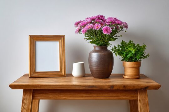 A rustic wooden table displays a golden picture frame a brown vase with pink chrysanthemums and a small potted plant - Powered by Adobe